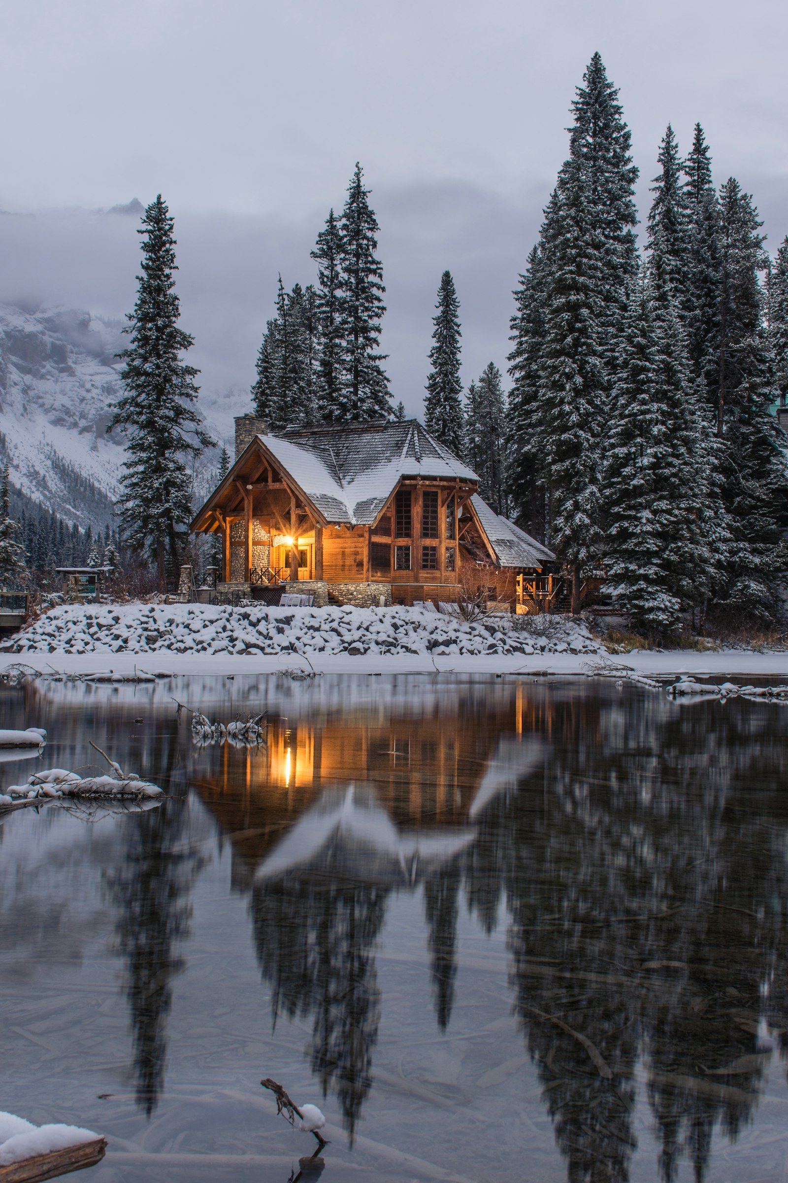 A wooden cabin beside a frozen pond and pines after snowfall. Photo by Ian Keefe on Unsplash.